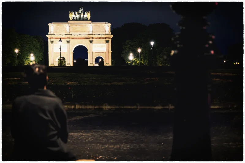 Thierry · 17/05/2026 · 3840 x 2560 Cette photographie met en scène un **arc de triomphe illuminé** la nuit, probablement l'Arc de Triomphe de Paris. Le **sujet principal** est cet arc, éclairé et dominant la composition. Le **cadrage** est serré sur une personne assise en premier plan, dont on ne voit que le dos, créant une perspective profonde.La **lumière** est douce et artificielle, mettant en valeur les contours de l'arc. Les **couleurs dominantes** sont des teintes sombres (noir, gris) avec des éclairages chauds (jaunes et blancs). L’**ambiance** est **nostalgique et contemplative**, renforcée par la nuit et les jeux d'ombres. Le style est **réaliste et photographique**, avec une touche poétique.
