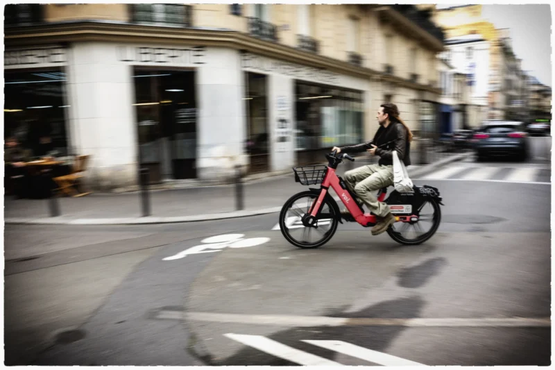 Thierry · 20/05/2026 · 3840 x 2560 Cette photographie capture un cycliste comme sujet principal, roulant sur un vélo cargo rouge. La composition montre une rue urbaine avec des bâtiments en arrière-plan légèrement floutés, suggérant un mouvement rapide. Le cadrage est serré sur le cycliste, centré dans l’image.La lumière est naturelle, probablement en journée, avec des tons doux et une ambiance légèrement contrastée. Les couleurs dominantes sont les teintes grises de la ville, rehaussées par le rouge vif du vélo. Le style est dynamique et réaliste, évoquant une ambiance moderne et urbaine, avec un flou artistique qui accentue le mouvement et l’énergie de la scène.