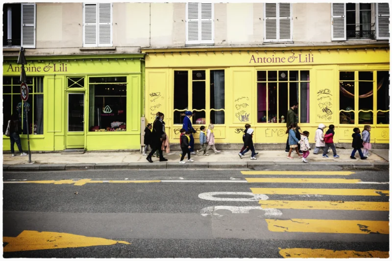 Thierry · 20/05/2026 · 3840 x 2560 Cette photographie capture une rue urbaine animée. Le sujet principal est un groupe de personnes marchant devant un bâtiment jaune vif, marqué "Annette & Lilit". La composition montre une perspective légèrement basse, avec des lignes directrices menant vers le bâtiment.Le cadrage est serré sur les passants et l’établissement. La lumière, naturelle et douce, semble tamisée, créant des ombres légères. Les couleurs dominantes sont le jaune vif du bâtiment et les tons neutres des vêtements et du bitume.Le style est réaliste et documentaire, évoquant une ambiance quotidienne et vivante dans un quartier urbain.