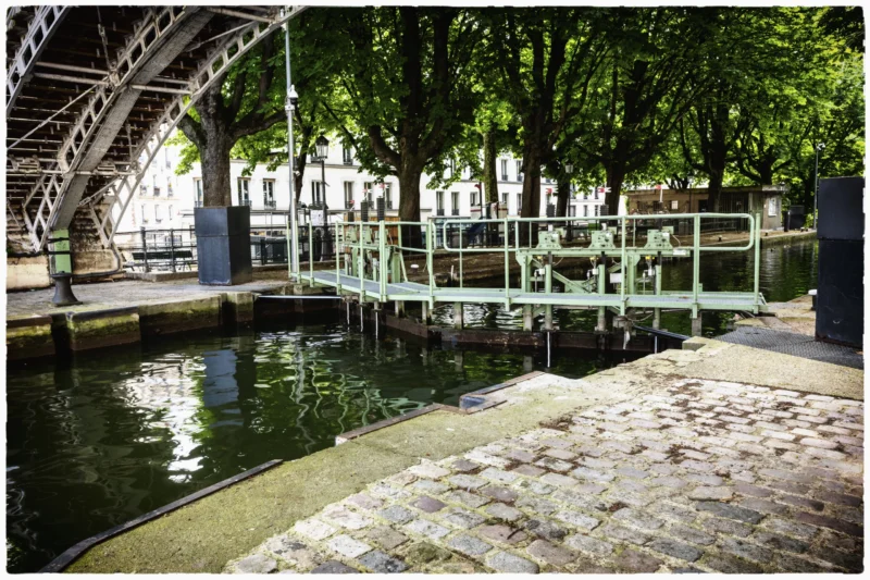 Thierry · 20/05/2026 · 3840 x 2560 Cette photographie montre un bassin d’eau calme sous une structure métallique en arc, évoquant un pont ou une passerelle. Le sujet principal est l’eau entourée de berges en pierre et de végétation luxuriante.La composition est équilibrée, avec des lignes horizontales et verticales créées par le bassin et les arbres. Le cadrage est serré, mettant en valeur les détails de l’eau et des structures environnantes. La lumière est naturelle et douce, suggérant un jour ensoleillé mais sans éblouissement. Les couleurs dominantes sont les verts des arbres, les gris de la pierre et du métal, et les bleus de l’eau.Le style est réaliste et photographique, avec une ambiance paisible et légèrement urbaine.