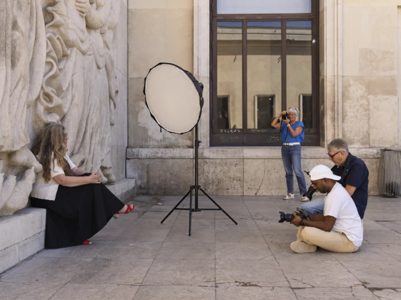 Cette photographie capture une scène de rue artistique. Le sujet principal semble être une séance photo improvisée : une personne assise, photographiée par un photographe équipé d’un flash reflété. La composition montre un cadrage serré sur les sujets, avec un arrière-plan architectural sobre.La lumière est naturelle, douce, avec des ombres marquées. Les couleurs dominantes sont neutres et naturelles : gris des murs, tons chauds des vêtements et teintes claires de la peau. Le style est documentaire et spontané, évoquant une ambiance artistique et détendue, presque intimiste malgré le cadre urbain.