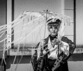 Taunia · 2200 x 1650 Cette photographie en noir et blanc met en scène un personnage principal vêtu d’un costume militaire et d’un chapeau, tenant un parapluie transparent rempli de fils blancs. La composition est centrée sur ce sujet, avec un cadrage serré qui accentue son expression et son accessoire. La lumière, douce et contrastée, souligne les détails du costume et crée des ombres marquées. Les tons dominants, en niveaux de gris, renforcent l’aspect dramatique et mystérieux. Le style évoque une touche artistique et surréaliste, conférant une ambiance énigmatique et théâtrale à l’image.
