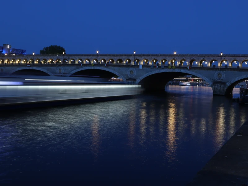 Francoise · 23/05/2026 · 6000 x 4000 Cette photographie capture un **bateau** en mouvement sous un **pont éclairé**, probablement un pont de Paris. Le **sujet principal** est le trajet lumineux du bateau, créant une traînée bleutée sur l’eau.La **composition** est dynamique, avec un **cadrage horizontal** mettant en valeur la perspective du pont et de l’eau. La **lumière** est douce, avec des éclairages artificiels soulignant les structures. Les **couleurs dominantes** sont les bleus profonds du ciel et de l’eau, contrastant avec les teintes chaudes des lumières.Le **style** est photographique moderne, avec une **ambiance nocturne sereine et légèrement mystérieuse**.