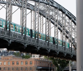 Claude · 5665 x 3777 Cette photographie montre un **train turquoise** traversant un **pont métallique** à structure complexe. Le sujet principal est le train en mouvement, encadré par les poutres du pont.La **composition** est dynamique, avec une perspective légèrement basse. Le **cadrage** isole partiellement le pont et le train, tout en incluant des bâtiments en arrière-plan. La **lumière** semble naturelle, diffuse, sans ombre marquée.Les **couleurs dominantes** sont les tons métalliques gris, le turquoise du train et les teintes chaudes des bâtiments. Le style est **documentaire et urbain**, évoquant une ambiance **industrielle et moderne**.