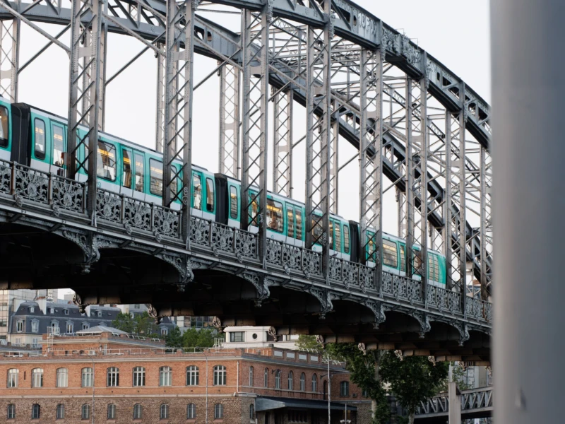 Claude · 5665 x 3777 Cette photographie montre un **train turquoise** traversant un **pont métallique** à structure complexe. Le sujet principal est le train en mouvement, encadré par les poutres du pont.La **composition** est dynamique, avec une perspective légèrement basse. Le **cadrage** isole partiellement le pont et le train, tout en incluant des bâtiments en arrière-plan. La **lumière** semble naturelle, diffuse, sans ombre marquée.Les **couleurs dominantes** sont les tons métalliques gris, le turquoise du train et les teintes chaudes des bâtiments. Le style est **documentaire et urbain**, évoquant une ambiance **industrielle et moderne**.