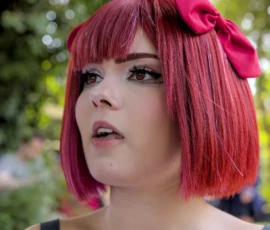 Thibault · 24/05/2026 · 1920 x 1595 Cette photographie met en scène une personne au regard expressif, avec une coiffure rose à boucles et un ruban assorti. Le sujet principal est centré, avec un cadrage serré sur son visage, isolant ainsi ses traits et son expression.La lumière semble naturelle, douce et légèrement tamisée, créant des ombres subtiles. Les couleurs dominantes sont les tons roses vifs de la chevelure et du ruban, contrastant avec la peau claire. L’ambiance évoque un style rétro ou inspiré des années 1960, avec une touche artistique et légèrement théâtrale.