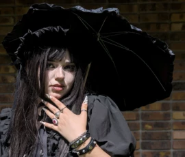 Thibault · 24/05/2026 · 1920 x 1371 Cette photographie met en scène une jeune femme comme sujet principal, vêtue d’une tenue sombre et élégante, avec un chapeau à larges bords et une longue écharpe. La composition est simple et centrée, avec un cadrage serré qui isole le visage et les épaules.La lumière est tamisée, créant des contrastes doux et des ombres marquées. Les couleurs dominantes sont les noirs et gris, renforçant une ambiance mystérieuse et gothique. Le style évoque un côté dramatique et romantique, accentué par le fond flou de briques, typique d’un décor urbain. L’ensemble dégage une atmosphère sombre et introspective.