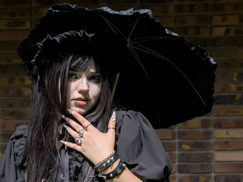 Thibault · 24/05/2026 · 1920 x 1371 Cette photographie met en scène une jeune femme comme sujet principal, vêtue d’une tenue sombre et élégante, avec un chapeau à larges bords et une longue écharpe. La composition est simple et centrée, avec un cadrage serré qui isole le visage et les épaules.La lumière est tamisée, créant des contrastes doux et des ombres marquées. Les couleurs dominantes sont les noirs et gris, renforçant une ambiance mystérieuse et gothique. Le style évoque un côté dramatique et romantique, accentué par le fond flou de briques, typique d’un décor urbain. L’ensemble dégage une atmosphère sombre et introspective.