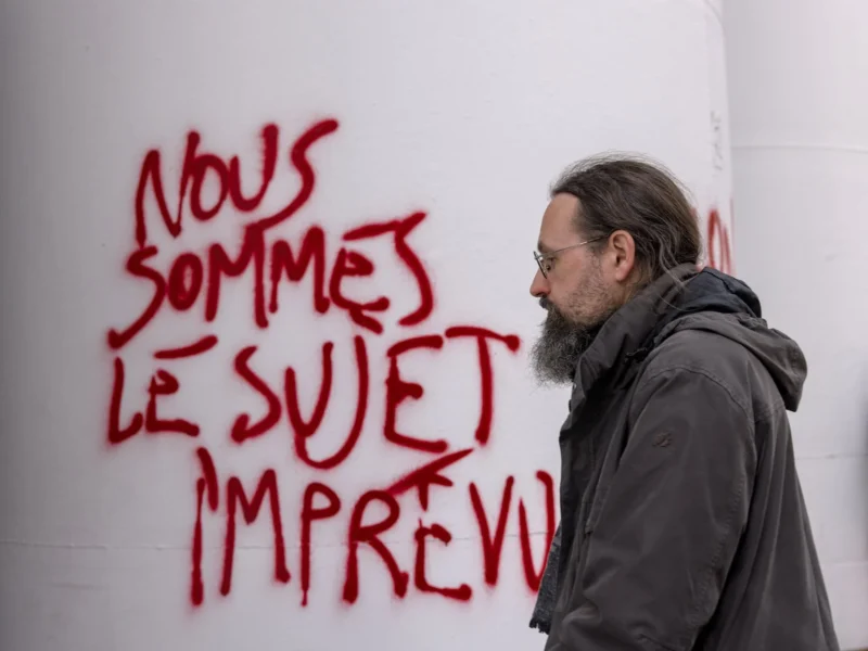 Thibault · 09/03/2025 · 1920 x 1440 Cette photographie montre un homme, sujet principal, observant un mur tagué. Le graffiti en rouge écrit *"Nous sommes le sujet imprévu"* attire l’attention. La composition est simple, avec un cadrage serré sur l’homme et le texte.La lumière semble naturelle, éclairant le sujet de face. Les couleurs dominantes sont le gris du mur, le rouge vif du graffiti et les tons neutres des vêtements de l’homme. Le style est documentaire, capturant un moment spontané. L’ambiance est à la fois contemplative et légèrement engagée, évoquant une réflexion sur un message social ou politique.
