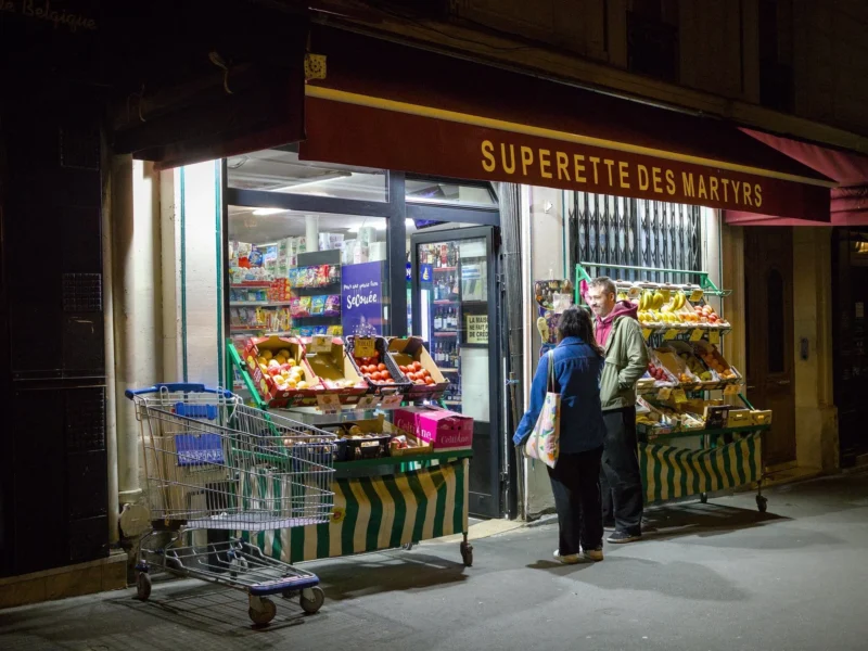 Thibault · 17/04/2025 · 1920 x 1440 Cette photographie montre une supérette de nuit, "Supérette des Marées". Le sujet principal est une cliente poussant un chariot rempli de produits, notamment des poissons et des fruits de mer.La composition met en valeur les produits bien éclairés à l'avant du magasin, avec une lumière artificielle chaude. Le cadrage est serré sur l'entrée, créant une ambiance intimiste. Les couleurs dominantes sont les tons froids des produits alimentaires et les éclairages chauds jaunes et blancs.Le style est réaliste et documentaire, capturant une scène quotidienne. L'ambiance est nocturne et vivante, malgré l'heure tardive.