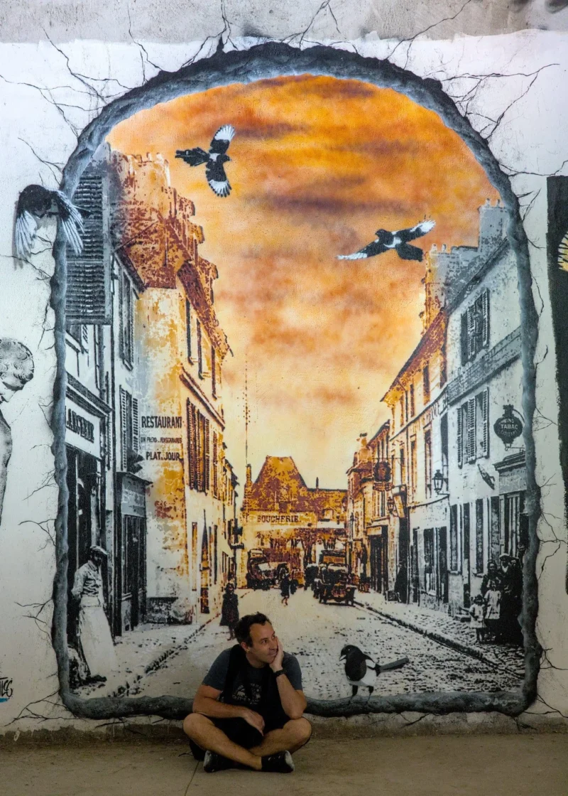 Thibault · 03/05/2025 · 1371 x 1920 Cette photographie montre un homme assis devant une fresque murale. Le sujet principal est cette œuvre street art représentant une rue urbaine avec des bâtiments et un ciel coloré.La composition est verticale, avec un cadrage serré sur le mur et le personnage. La lumière semble naturelle, éclairant le mur de manière homogène. Les couleurs dominantes sont les tons chauds (oranges, jaunes) contrastant avec les gris et noirs des bâtiments.Le style est réaliste et détaillé, évoquant une ambiance nostalgique et artistique, mêlant urbanité et créativité.