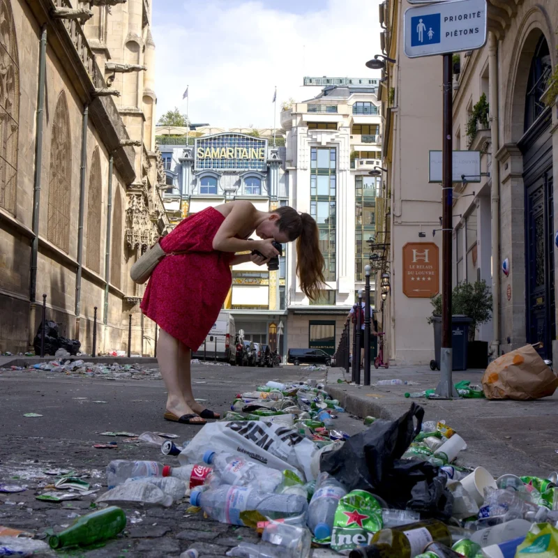 Thibault · 22/06/2025 · 1920 x 1920 Cette photographie montre une femme en robe rouge, sujet principal, penchée pour ramasser des déchets dans une rue encombrée. La composition met en contraste son action avec l’environnement négligé.Le cadrage est serré sur elle, avec une perspective légèrement basse, soulignant son effort. La lumière est naturelle, probablement en journée, avec des ombres marquées.Les couleurs dominantes sont les tons froids des déchets (blanc, gris) et le rouge vif de la robe, créant un contraste saisissant. Le style est documentaire et réaliste, évoquant une ambiance de détresse et d’urgence environnementale dans un cadre urbain.