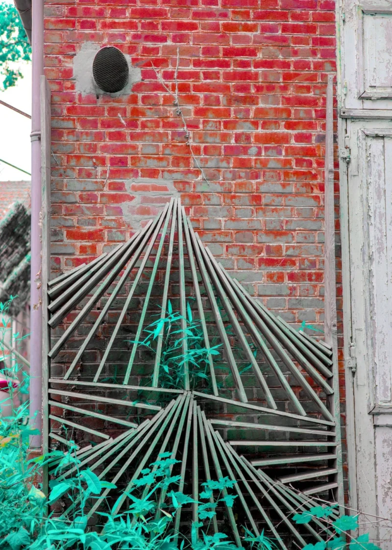 Thibault · 19/07/2025 · 1371 x 1920 Cette photographie met en scène un mur de briques rouges avec une structure métallique en forme d’éventail, partiellement cachée par des plantes vertes grimpantes. Le sujet principal est cette installation métallique, qui crée un contraste géométrique avec le mur organique.Le cadrage est serré, centré sur l’élément métallique. La lumière naturelle éclaire la scène, accentuant les textures et les ombres. Les couleurs dominantes sont le rouge des briques, le vert des plantes et le gris métallique. Le style est à la fois urbain et artistique, évoquant une ambiance à la fois industrielle et naturelle, presque poétique.