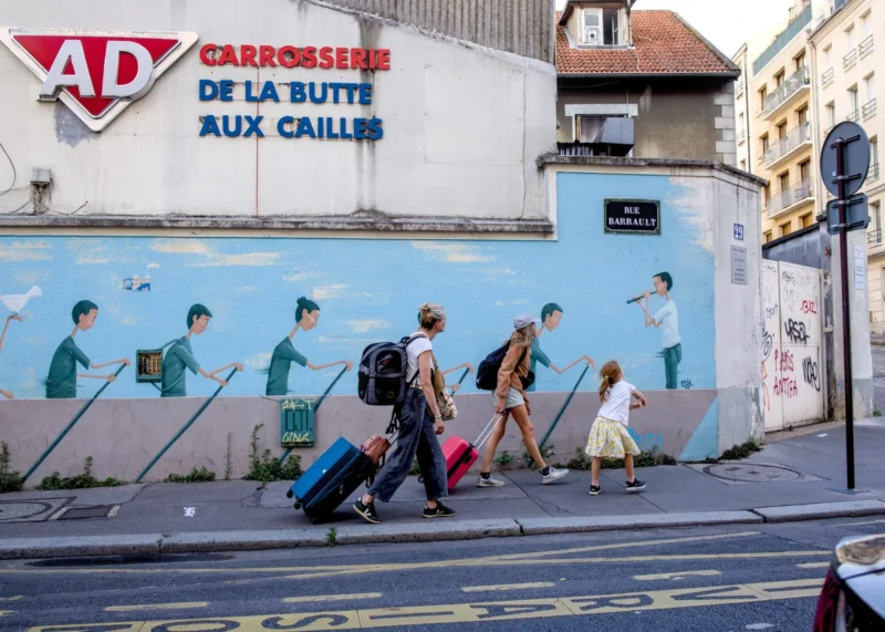 Thibault · 30/07/2025 · 1920 x 1371 Cette photographie montre des voyageurs marchant avec des bagages devant un mur mural. Le sujet principal est un groupe de personnes en mouvement, dont une enfant en robe blanche.La composition est dynamique, avec un cadrage serré sur les personnages et un arrière-plan urbain. La lumière est naturelle, probablement en journée, éclairant bien la scène. Les couleurs dominantes sont les tons bleus du mur, les gris de la ville et les couleurs vives des vêtements des passants.Le style est documentaire et réaliste, capturant une scène de vie quotidienne. L’ambiance est active et légèrement touristique, évoquant un lieu de transit comme une gare.