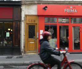 Marine · 16/05/2026 · 4000 x 3000 Cette photographie met en scène un cycliste en premier plan, flou, suggérant un mouvement. Le sujet principal secondaire est un bâtiment urbain avec deux enseignes : "LCB" et "PRIMA".La composition joue sur la profondeur de champ, opposant netteté et flou. Le cadrage est serré, centré sur les façades et le cycliste.Les couleurs dominantes sont les rouges (bâtiment, vélo) et les tons neutres (brique, bois). La lumière semble naturelle, probablement en journée, avec des ombres douces.L’ambiance est urbaine et dynamique, évoquant une ville animée. Le style est réaliste et documentaire.
