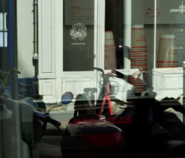 Marine · 16/05/2026 · 4000 x 3000 Cette photographie montre principalement un scooter garé devant une vitrine. La composition est simple, avec un cadrage serré sur le véhicule et son reflet dans la vitre. La lumière est tamisée, créant une ambiance peu lumineuse et légèrement floue.Les couleurs dominantes sont des tons neutres (gris, blanc) avec des touches de rouge et noir sur le scooter. Le style est réaliste et documentaire, sans mise en scène particulière. L’ambiance est urbaine et un peu terne, évoquant un lieu intérieur ou semi-clos, comme un magasin ou un hall.