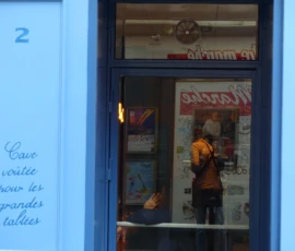 Marine · 16/05/2026 · 4000 x 3000 Cette photographie montre une vitrine de cave à vin avec une inscription manuscrite : *« Cave voilée pour les grandes tablées »*. Le sujet principal est une silhouette floue à l'intérieur, éclairée par une lumière chaude.La composition est symétrique, centrée sur la porte vitrée. Le cadrage est serré, mettant en valeur les détails de la vitrine et les inscriptions. Les couleurs dominantes sont les bleus froids de la porte et les tons chauds de la lumière intérieure. L’ambiance est intimiste et légèrement mystérieuse, évoquant un lieu chaleureux et convivial. Le style est réaliste et photographique, avec une touche poétique.