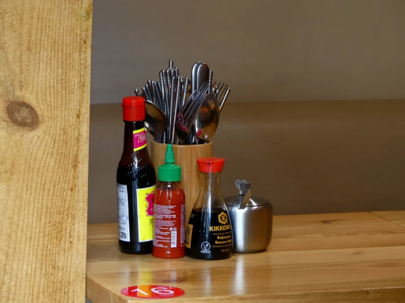 Marine · 16/05/2026 · 4000 x 3000 Cette photographie montre un ensemble d’objets posés sur un plan en bois clair. Le sujet principal est un assortiment de couverts (fourchettes et cuillères) dans un pot en verre, accompagné de bouteilles de condiments (sauces) et d’un petit saladier métallique.La composition est simple et fonctionnelle, avec un cadrage serré mettant en valeur les détails. La lumière naturelle, douce et légèrement tamisée, crée des ombres douces. Les couleurs dominantes sont les tons chauds (bois, rouge des étiquettes) et métalliques (argenté des couverts).Le style est épuré et minimaliste, évoquant une ambiance chaleureuse et domestique.