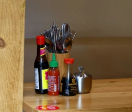 Marine · 16/05/2026 · 4000 x 3000 Cette photographie montre un ensemble d’objets posés sur un plan en bois clair. Le sujet principal est un assortiment de couverts (fourchettes et cuillères) dans un pot en verre, accompagné de bouteilles de condiments (sauces) et d’un petit saladier métallique.La composition est simple et fonctionnelle, avec un cadrage serré mettant en valeur les détails. La lumière naturelle, douce et légèrement tamisée, crée des ombres douces. Les couleurs dominantes sont les tons chauds (bois, rouge des étiquettes) et métalliques (argenté des couverts).Le style est épuré et minimaliste, évoquant une ambiance chaleureuse et domestique.