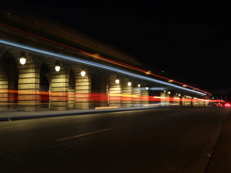 Francoise · 23/05/2026 · 6000 x 4000 Cette photographie capture une scène nocturne urbaine avec un **sujet principal** : les traînées lumineuses de véhicules en mouvement. La **composition** est horizontale, mettant en valeur une longue perspective de trottoir et de bâtiments. Le **cadrage** est serré sur la voie, créant un effet de profondeur.Les **lumières** dominent avec des teintes bleutées et rouges, typiques des phares et feux de voiture. Les **couleurs dominantes** sont sombres, avec des éclats lumineux vifs.Le **style** est dynamique, presque abstrait grâce aux traînées lumineuses. L’**ambiance** est nocturne et énergique, évoquant vitesse et mouvement dans une ville endormie.