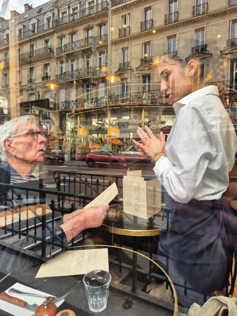 Bao · 3000 x 4000 Cette photographie montre deux hommes assis à une terrasse de café en extérieur. Le sujet principal est leur interaction, l’un lisant un livre tandis que l’autre semble discuter ou écouter.La composition est symétrique et naturelle, avec un cadrage serré sur les deux hommes. L’arrière-plan révèle des bâtiments parisiens typiques, ajoutant du contexte urbain.La lumière est douce et naturelle, probablement en journée. Les couleurs dominantes sont neutres et chaudes, avec des tons beiges, gris et blancs, rehaussés par des touches de vert et de bleu.L’ambiance est calme et conviviale, évoquant une pause détendue en ville.