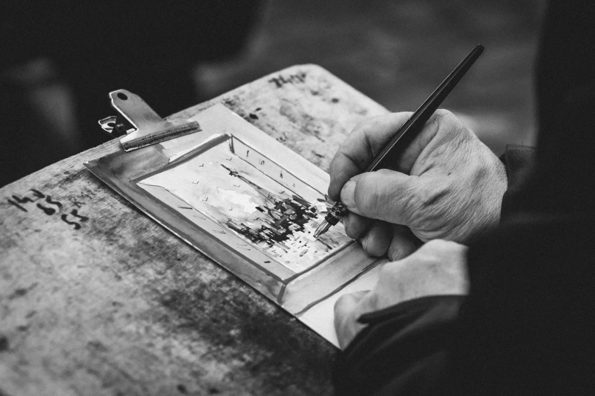 Marc L. · 21/03/2026 · 3589 x 2393 Cette photographie en noir et blanc met en scène une personne en train de dessiner ou d'écrire dans un carnet ouvert. Le sujet principal est la main tenant un stylo, ainsi que le carnet posé sur une surface.La composition est centrée sur les détails précis de l'action, avec un cadrage serré. La lumière douce et contrastée accentue les textures et les ombres, créant une ambiance intime et studieuse. Les tons dominants sont les gris et noirs, renforçant une atmosphère classique et contemplative. Le style évoque un travail artisanal et minutieux, évoquant concentration et créativité.