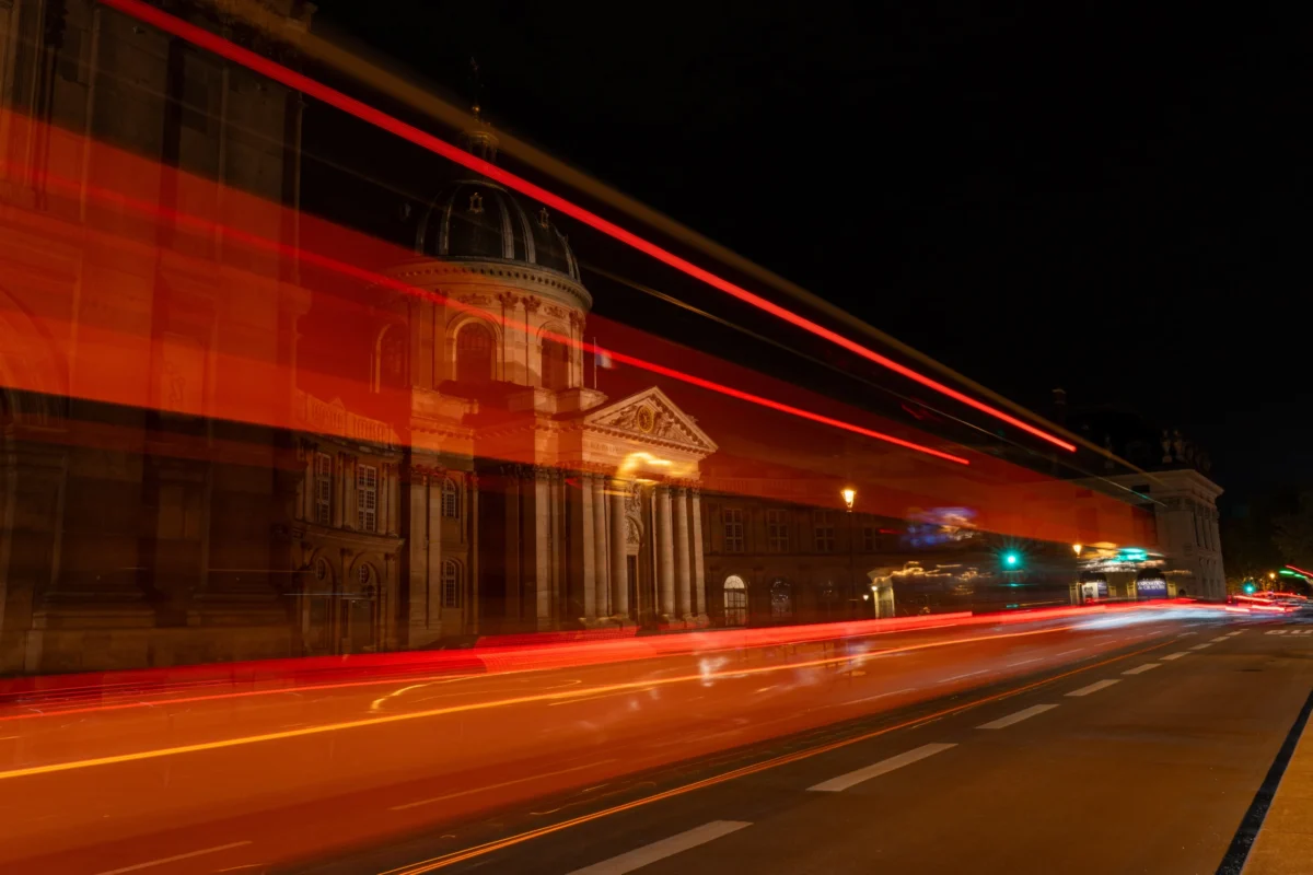 Marc L. · 10/04/2026 · 7008 x 4672 Cette photographie capture une scène nocturne urbaine avec un **sujet principal** axé sur les **traînées lumineuses** des phares et feux de véhicules en mouvement. La **composition** met en valeur une architecture classique éclairée, avec des lignes horizontales et verticales bien définies.Le **cadrage** est serré sur un bâtiment et une rue, créant une perspective dynamique. Les **lumières** sont vives et contrastées, avec des **couleurs dominantes** chaudes (rouges, oranges) et des teintes froides (bleues des néons). Le **style** relève de la photographie longue exposition, donnant un effet de vitesse et de mouvement.L’**ambiance** est à la fois moderne et nostalgique, évoquant l’énergie vibrante d’une ville la nuit.
