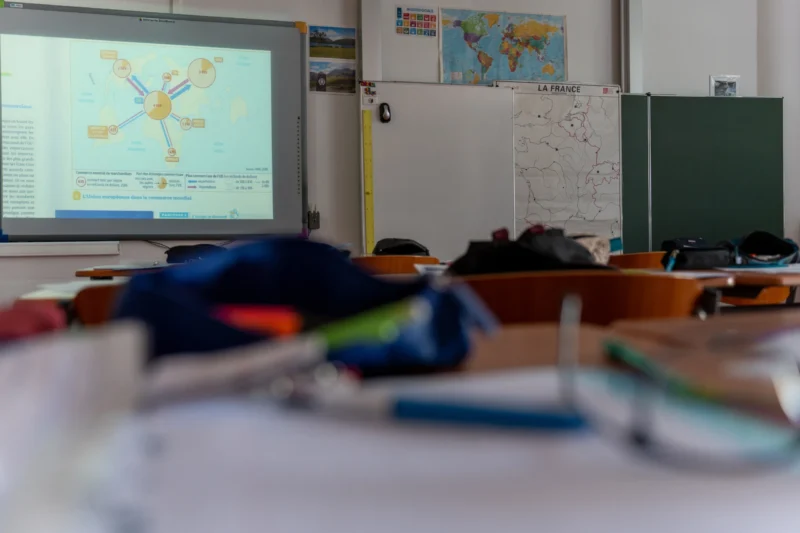Marc L. · 26/05/2026 · 6771 x 4514 Cette photographie montre principalement une **classe scolaire**. Le sujet central est un **écran de projection** affichant un schéma, avec des tables et des chaises en premier plan légèrement floues.La **composition** est dynamique, avec un cadrage serré sur le bureau avant-plan. La **lumière** semble naturelle et douce, éclairant uniformément la scène. Les **couleurs dominantes** sont neutres (beige, gris) avec des touches de bleu et vert (affiches, vêtements).Le style est **documentaire et réaliste**, capturant un environnement quotidien. L’**ambiance** est studieuse et un peu désordonnée, typique d’une salle de classe en activité.