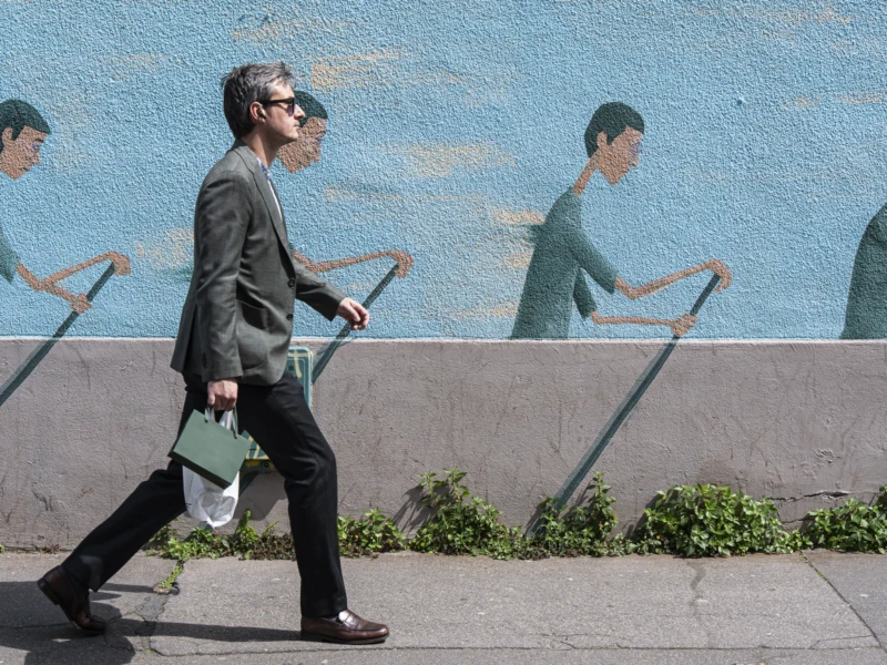 Jean-Yves · 09/05/2026 · 3000 x 1687 Cette photographie montre une personne marchant devant un mur mural. Le **sujet principal** est cette personne, vêtue d’un manteau sombre, avec des lunettes et une sacoche.La **composition** joue sur le contraste entre le mouvement réel de la personne et les silhouettes statiques du mural, répétées en miroir. Le **cadrage** est serré sur le sujet, avec un arrière-plan partiel.La **lumière** est naturelle et douce, éclairant les tons clairs du mural. Les **couleurs dominantes** sont les bleus du fond et les gris de la personne et du sol. Le style est **urbain et graphique**, créant une **ambiance** à la fois dynamique et contemplative.