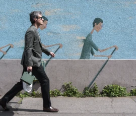 Jean-Yves · 09/05/2026 · 3000 x 1687 Cette photographie montre une personne marchant devant un mur mural. Le **sujet principal** est cette personne, vêtue d’un manteau sombre, avec des lunettes et une sacoche.La **composition** joue sur le contraste entre le mouvement réel de la personne et les silhouettes statiques du mural, répétées en miroir. Le **cadrage** est serré sur le sujet, avec un arrière-plan partiel.La **lumière** est naturelle et douce, éclairant les tons clairs du mural. Les **couleurs dominantes** sont les bleus du fond et les gris de la personne et du sol. Le style est **urbain et graphique**, créant une **ambiance** à la fois dynamique et contemplative.