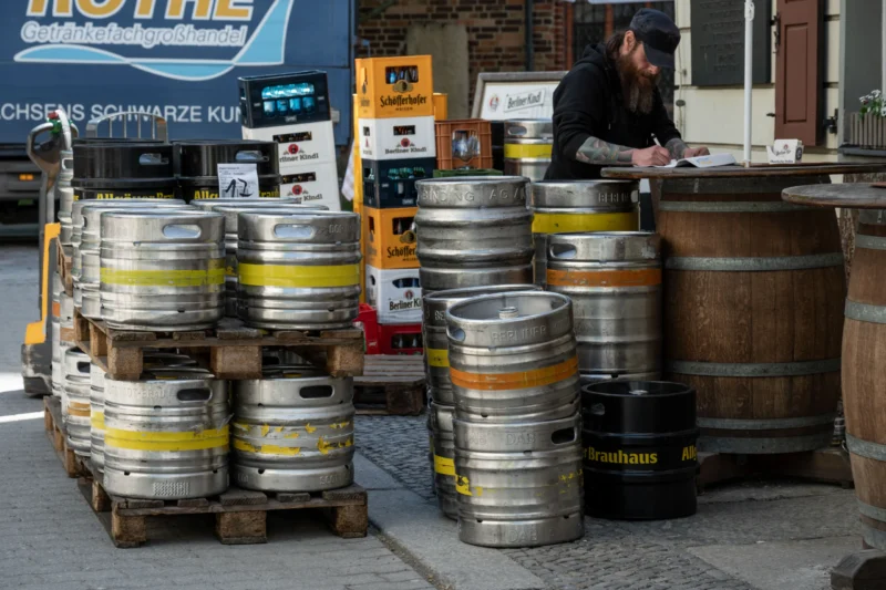 Jean-Marc · 30/04/2026 · 5489 x 3659 Cette photographie montre principalement des **fûts de bière** empilés sur des palettes, avec quelques barils en bois. La composition met en scène un **marché ou une vente de produits brassicoles**, avec une personne en arrière-plan, probablement en train de gérer les stocks.Le **cadrage** est serré sur les fûts, avec une légère profondeur de champ. La **lumière** semble naturelle, légèrement tamisée, créant des ombres douces. Les **couleurs dominantes** sont les tons métalliques (argent, jaune) des fûts, contrastant avec le bois et les teintes neutres du sol.L’ambiance est **artisanale et professionnelle**, évoquant un commerce spécialisé dans les boissons. Le style est **documentaire et réaliste**.