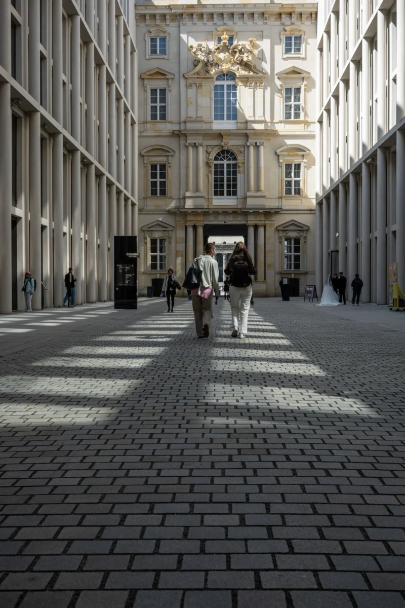 Jean-Marc · 30/04/2026 · 6048 x 4032 Cette photographie capture une cour pavée urbaine avec des bâtiments classiques en arrière-plan. Le sujet principal est une personne poussant un vélo, accompagnée d’une autre silhouette. La composition est symétrique, avec des lignes directrices convergeant vers le fond.Le cadrage est serré, mettant en valeur les détails architecturaux et les jeux d’ombre. La lumière naturelle, douce et légèrement rasante, crée des contrastes subtils. Les couleurs dominantes sont les tons gris-beige des pavés et des bâtiments, adoucis par des touches claires.Le style est épuré et réaliste, évoquant une ambiance calme et introspective, presque mélancolique.
