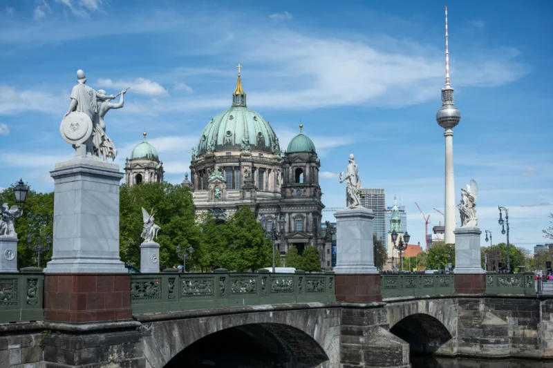 Jean-Marc · 30/04/2026 · 6048 x 4032 Cette photographie montre un pont orné de statues, avec en arrière-plan le **dôme doré de la cathédrale Saint-Hedwige** et la **tour de télévision de Berlin (Fernsehturm)**. La composition met en valeur ces éléments architecturaux emblématiques.Le cadrage est large, capturant une vue panoramique. La lumière est naturelle, éclatante, avec un ciel dégagé. Les couleurs dominantes sont les tons **beige, vert et bleu**, adoucis par les ombres. Le style est réaliste et photographique, évoquant une ambiance **urbaine majestueuse et historique**.