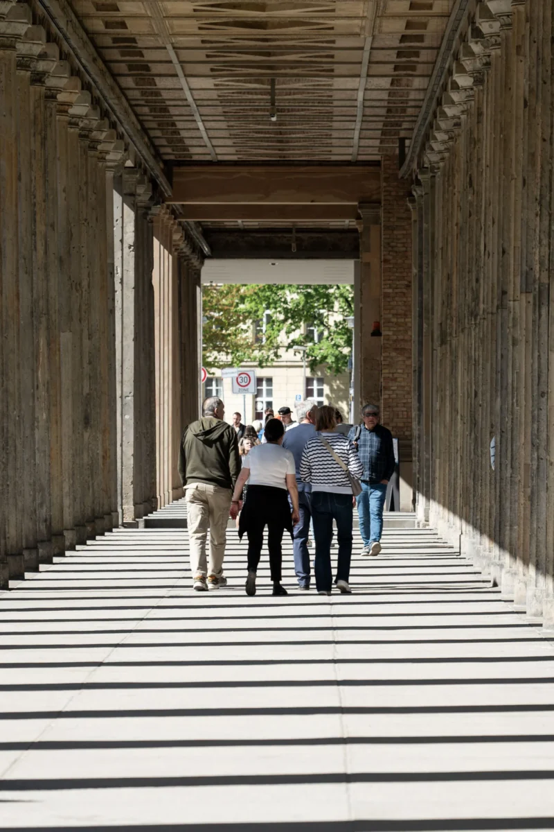 Jean-Marc · 01/05/2026 · 3164 x 2109 Cette photographie montre un groupe de personnes marchant sous une passerelle moderne. Le **sujet principal** est l’architecture et les passants en mouvement.La **composition** est dynamique, avec une perspective axiale menant vers la lumière naturelle en fond. Le **cadrage** est serré, mettant en valeur les lignes horizontales et verticales des structures en béton.La **lumière** est naturelle et douce, créant des ombres nettes. Les **couleurs dominantes** sont les tons gris-beige du béton et les verts des arbres en arrière-plan.Le style est épuré et contemporain, évoquant une **ambiance** à la fois urbaine et apaisante.
