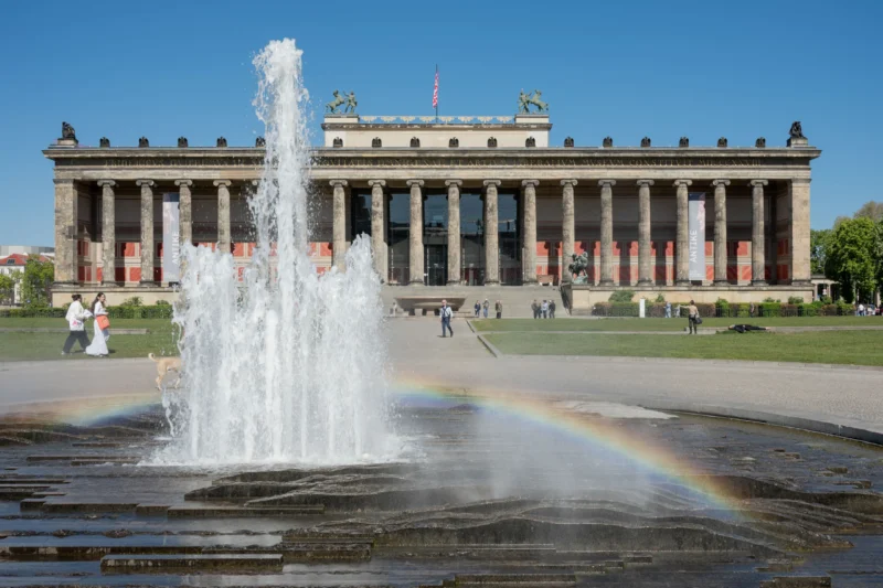 Jean-Marc · 01/05/2026 · 5309 x 3539 Cette photographie met en scène un **fountain** en premier plan avec un **arc-en-ciel** formé par la lumière du soleil. En arrière-plan, un **grand bâtiment néoclassique** (l'Alte Nationalgalerie de Berlin) domine la composition. Le cadrage est large, intégrant des éléments naturels et architecturaux.La lumière est **vive et naturelle**, créant des contrastes nets. Les **couleurs dominantes** sont les tons pastel du bâtiment (beige, gris), le vert des arbres et le bleu du ciel. L’ambiance est **sereine et majestueuse**, mêlant harmonie naturelle et grandeur architecturale. Le style est **réaliste et photographique**, mettant en valeur la beauté classique et la lumière.