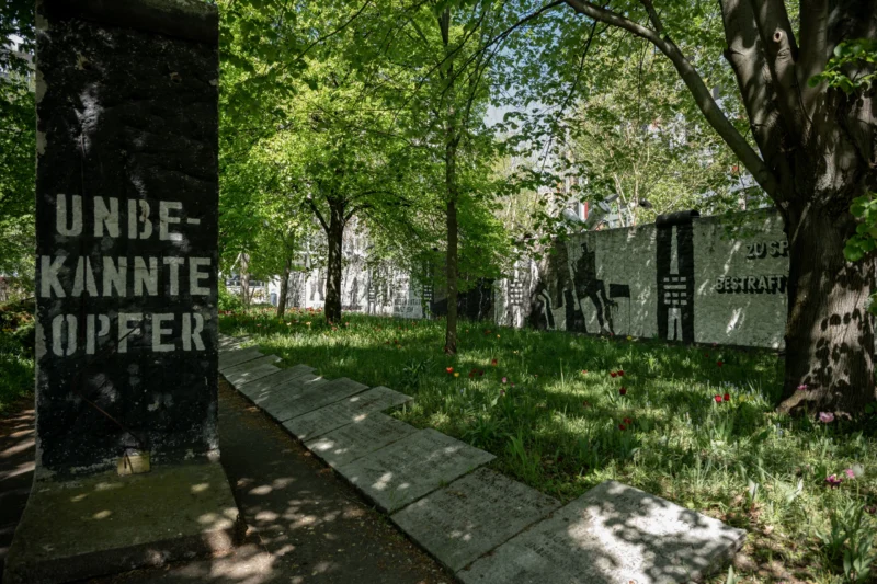 Jean-Marc · 01/05/2026 · 6048 x 4032 Cette photographie montre un mémorial dédié aux victimes inconnues, marqué par une stèle noire avec l'inscription *« UNBEKANNTE OPFER »*. La composition met en valeur une allée pavée bordée d'arbres et de végétation verdoyante, menant vers un bâtiment gris en arrière-plan.Le cadrage est serré sur la stèle, avec une perspective légèrement oblique. La lumière naturelle, douce et tamisée, crée une ambiance calme et solennelle. Les couleurs dominantes sont les verts des arbres et le gris des pavés et du bâtiment, renforçant une atmosphère sobre et mélancolique, typique des lieux commémoratifs.