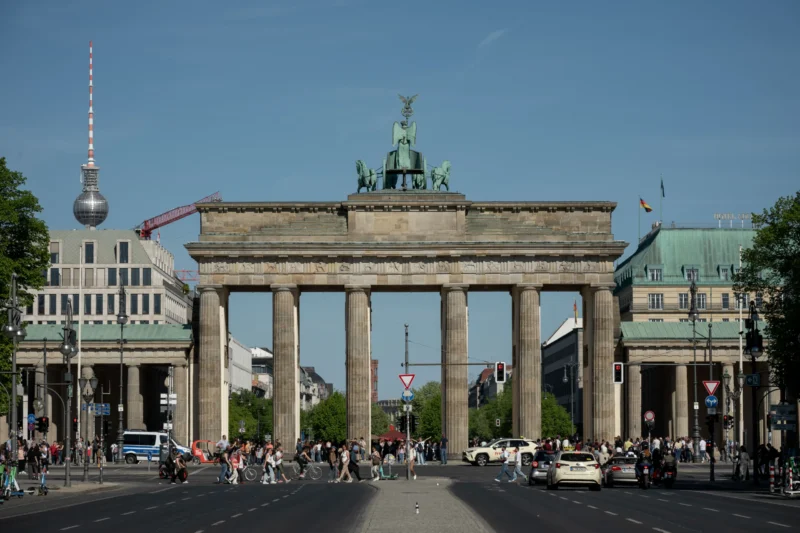 Jean-Marc · 01/05/2026 · 5951 x 3967 Cette photographie met en scène la **porte de Brandebourg** à Berlin, sujet principal emblématique. La composition est symétrique, centrée sur l'arc de triomphe avec ses colonnes et la quadrige en son sommet. Le cadrage inclut une avenue animée et des bâtiments environnants.La lumière est naturelle, éclatante, suggérant un jour ensoleillé. Les couleurs dominantes sont les tons gris-beige des bâtiments, contrastés par le vert des arbres et le bleu du ciel. Le style est documentaire, réaliste, capturant une scène urbaine dynamique. L’ambiance est à la fois historique et moderne, reflétant l’énergie de Berlin.
