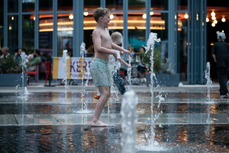 Jean-Marc · 01/05/2026 · 6048 x 4032 Cette photographie met en scène deux enfants jouant dans une fontaine urbaine. Le sujet principal est leur interaction joyeuse avec les jets d’eau.La composition est dynamique, centrée sur les enfants, avec un cadrage serré qui isole leur activité. L’arrière-plan flou montre des bâtiments et des lumières, créant une ambiance nocturne.La lumière est douce et tamisée, avec des éclairages artificiels qui adoucissent les contours. Les couleurs dominantes sont des tons froids (bleu nuit, gris) contrastés par les éclats de lumière et les vêtements colorés des enfants.Le style est naturel et spontané, évoquant une ambiance ludique et détendue.