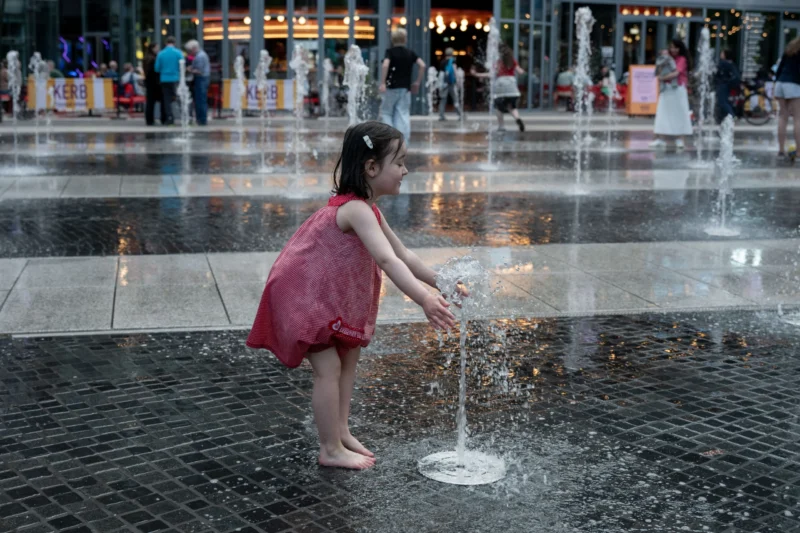 Jean-Marc · 01/05/2026 · 5522 x 3681 Cette photographie met en scène une jeune fille en robe rose comme sujet principal, jouant dans un jet d’eau. La composition est dynamique, centrée sur l’enfant, avec un cadrage serré qui isole son action. L’arrière-plan flou montre des fontaines et des passants, créant une ambiance urbaine animée.La lumière est naturelle, douce et légèrement tamisée, probablement en journée. Les couleurs dominantes sont les tons pastel (rose, gris) et les éclats d’eau scintillante. Le style est documentaire et spontané, capturant un moment authentique et joyeux. L’ambiance est à la fois ludique et sereine.