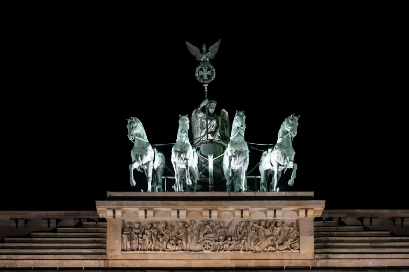Jean-Marc · 01/05/2026 · 5728 x 3819 Cette photographie met en scène la **Quadrige de Berlin**, char tiré par quatre chevaux, surmonté d’une **aigle**. La composition est symétrique, centrée sur le monument. Le cadrage serré isole la structure, avec un arrière-plan noir qui accentue les détails.La lumière, douce et contrastée, souligne les contours métalliques et les reliefs sculptés. Les **couleurs dominantes** sont les teintes vert-bleutées du bronze et les tons chauds du socle en pierre.Le style est **classique et monumental**, évoquant une ambiance solennelle et historique, renforcée par l’éclairage nocturne dramatique.