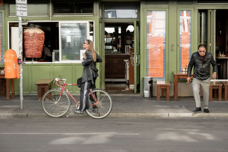 Jean-Marc · 02/05/2026 · 5367 x 3578 Cette photographie capture une scène urbaine avec deux hommes comme sujets principaux. L’un, près d’un vélo rouge, semble discuter avec une personne à l’intérieur d’un local vert, tandis que l’autre, en arrière-plan, observe.La composition est équilibrée, avec un cadrage serré sur les personnages et l’architecture. La lumière est naturelle, douce et légèrement tamisée, créant une ambiance calme. Les couleurs dominantes sont les tons verts (bâtiment), gris (asphalte) et rouge (vélo), avec des touches de brun et de beige.Le style est réaliste et documentaire, évoquant une ambiance quotidienne et authentique dans un cadre urbain paisible.