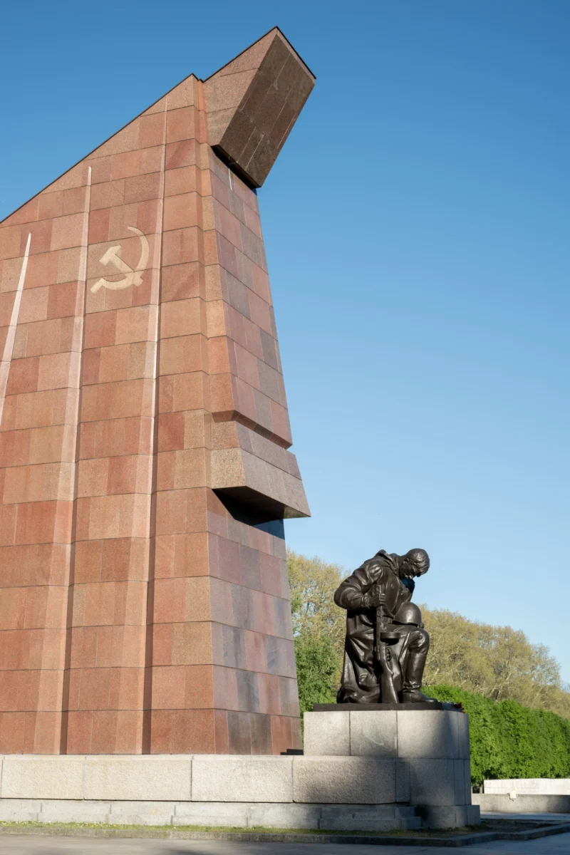Jean-Marc · 02/05/2026 · 5721 x 3814 Cette photographie montre un monument commémoratif. Le sujet principal est une statue représentant deux soldats en position de repos ou de soutien mutuel, devant une structure murale imposante en pierre aux teintes rougeâtres. Le symbole communiste (faucille et marteau) est gravé sur le mur.La composition est symétrique, avec un cadrage serré sur la statue et le mur. La lumière est naturelle, éclairant uniformément la scène sous un ciel bleu. Les couleurs dominantes sont les tons rouges et bruns du mur, contrastant avec le gris de la statue. L’ambiance est solennelle et mémorielle, évoquant le sacrifice et l’histoire.