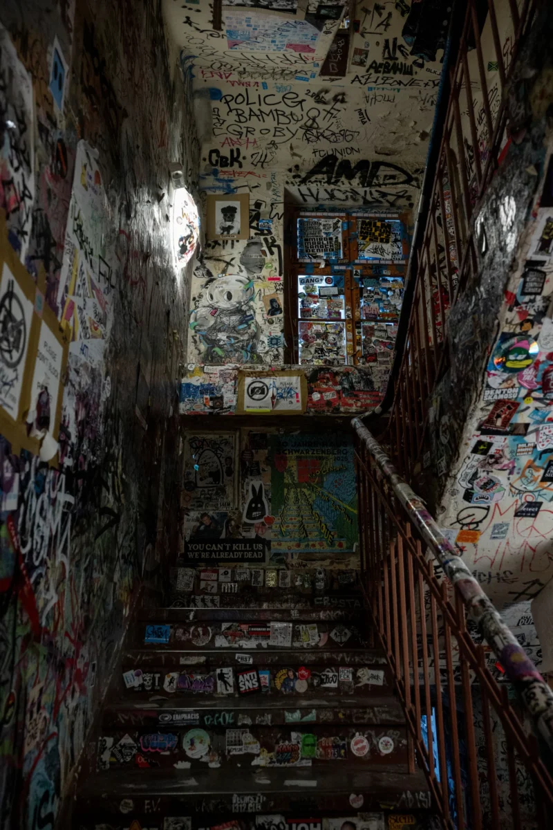 Jean-Marc · 02/05/2026 · 6048 x 4032 Cette photographie montre un escalier étroit et sombre recouvert de nombreuses graffitis et stickers. Le sujet principal est cet escalier, dont les murs et les marches sont ornés de dessins variés et colorés.La composition est verticale, avec un cadrage serré qui accentue la hauteur et l'étroitesse de l'espace. La lumière, tamisée et artificielle, crée une ambiance mystérieuse et intimiste. Les couleurs dominantes sont des teintes vives et contrastées, mais aussi des tons sombres, renforçant l'atmosphère unique et artistique du lieu. Le style évoque un mélange de street art et de décoration improvisée, donnant une impression de créativité et de vie urbaine.