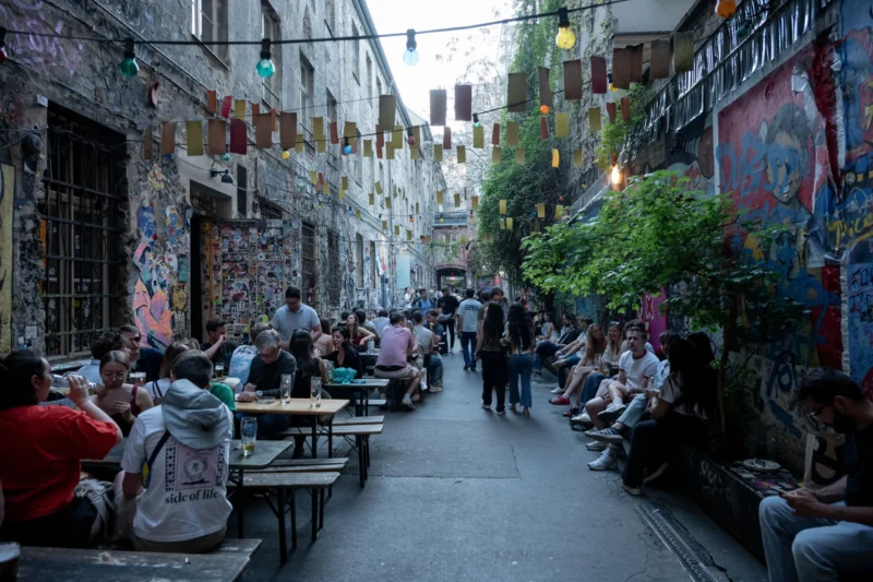 Jean-Marc · 02/05/2026 · 6048 x 4032 Cette photographie capture une ruelle animée, probablement un quartier artistique. Le sujet principal est un groupe de personnes assises à des tables en plein air, discutant ou prenant un repas. La composition montre une perspective en profondeur, avec des murs couverts de graffitis et des lanternes suspendues.Le cadrage est serré, mettant en valeur les détails des visages et des décorations murales. La lumière est douce, avec des ombres marquées, suggérant un coucher de soleil. Les couleurs dominantes sont des tons chauds (ocre, rouge) et des contrastes froids (bleus, verts des graffitis). L’ambiance est conviviale et artistique, typique d’un lieu culturel et vivant.
