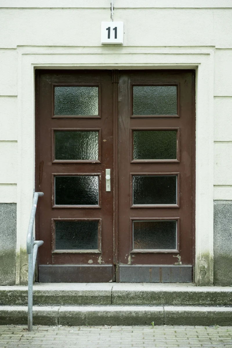 Jean-Marc · 03/05/2026 · 5836 x 3891 Cette photographie montre une porte d'entrée en bois sombre, numérotée 11, encastrée dans un mur blanc. Le sujet principal est cette porte à panneaux vitrés, avec une petite marche et un garde-corps métallique.La composition est symétrique et épurée, centrée sur la porte. Le cadrage est serré, mettant en valeur les détails architecturaux. La lumière semble naturelle et douce, créant des contrastes subtils entre les ombres et les surfaces claires. Les couleurs dominantes sont le blanc du mur, le brun foncé de la porte et le gris des pavés.L'ambiance est sobre et minimaliste, évoquant une élégance intemporelle.