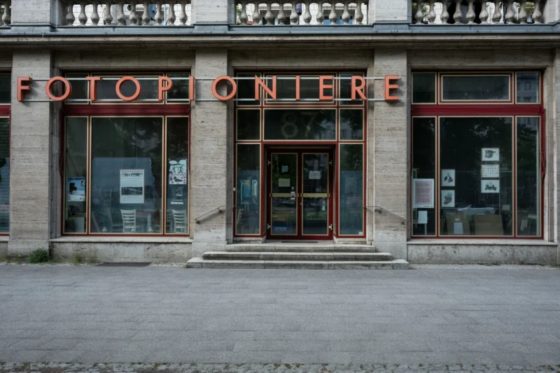 Jean-Marc · 03/05/2026 · 5776 x 3851 Cette photographie montre la façade d’un ancien magasin nommé **"Fotopionier"**. Le sujet principal est l’entrée vitrée et les enseignes en lettres métalliques rouges.La composition est symétrique, avec un cadrage serré sur la boutique. La lumière naturelle, douce et légèrement tamisée, crée des ombres discrètes. Les couleurs dominantes sont les tons beiges des pierres, le rouge des enseignes et le gris des vitres.Le style évoque un charme rétro et industriel, renforcé par les détails architecturaux. L’ambiance est calme et nostalgique, évoquant une époque passée.