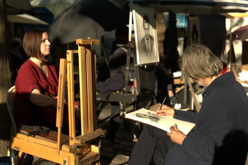 Bao · 6024 x 4016 Cette photographie montre deux personnes en train de dessiner ou peindre. Le sujet principal est l’activité artistique, avec une personne utilisant une grande toile sur un chevalet et l’autre dessinant sur une feuille.La composition est dynamique, avec un cadrage serré sur les artistes. La lumière semble naturelle et tamisée, créant des ombres douces. Les couleurs dominantes sont des tons neutres et chauds, avec des touches de rouge et de bleu.L’ambiance est studieuse et artistique, évoquant un atelier ou un espace créatif. Le style semble réaliste et attentif aux détails.