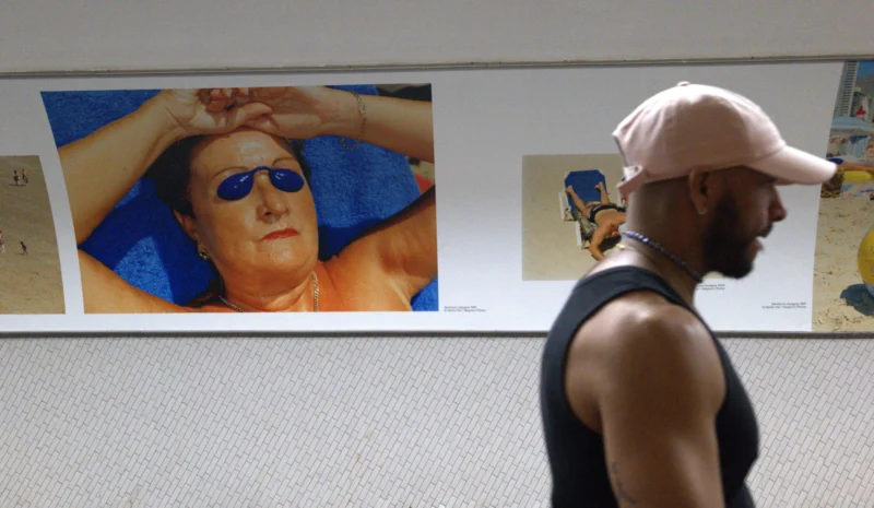 Bao · 5737 x 3337 Cette photographie montre un homme en mouvement, vêtu d’un chapeau et d’un tank top, passant devant une affiche publicitaire. Le sujet principal est cette affiche représentant un visage masculin stylisé, aux traits exagérés et aux couleurs vives.La composition oppose le mouvement du personnage à la fixité de l’affiche. Le cadrage est serré sur les deux éléments, avec une lumière naturelle et diffuse. Les couleurs dominantes sont vives et contrastées (bleu, rose, jaune) pour l’affiche, tandis que l’homme est dans des tons neutres.L’ambiance est à la fois urbaine et artistique, mêlant quotidien et esthétique pop.