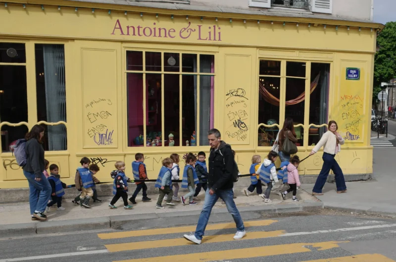 Bao · 3448 x 2279 Cette photographie montre une rue animée devant un café-restaurant nommé **« Antoine & Lili »**. Le sujet principal est un groupe d’enfants jouant devant l’établissement, encadrés par un adulte.La composition est dynamique, avec un cadrage large capturant la vie urbaine. Les couleurs dominantes sont les tons chauds (jaune du bâtiment, rouge des enseignes) contrastant avec les gris de la ville. La lumière est naturelle, probablement en journée, éclairant la scène de manière homogène.L’ambiance est joyeuse et vivante, reflétant un moment de détente et de sociabilité en ville. Le style est documentaire et spontané.