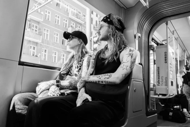 Alice · 03/05/2026 · 7008 x 4672 Cette photographie en noir et blanc met en scène deux personnes assises dans un train, l’une dormant ou détendue, l’autre éveillée et les observant. Le sujet principal est cette interaction silencieuse et intime.La composition joue sur le contraste entre les deux personnages et l’architecture urbaine vue par la fenêtre. Le cadrage est serré, centré sur eux, avec une profondeur de champ réduite.La lumière est douce et naturelle, filtrée par les vitres, créant une ambiance calme et mélancolique. Les tons dominants sont les gris et noirs, renforçant l’aspect intemporel et contemplatif. Le style est documentaire et poétique.